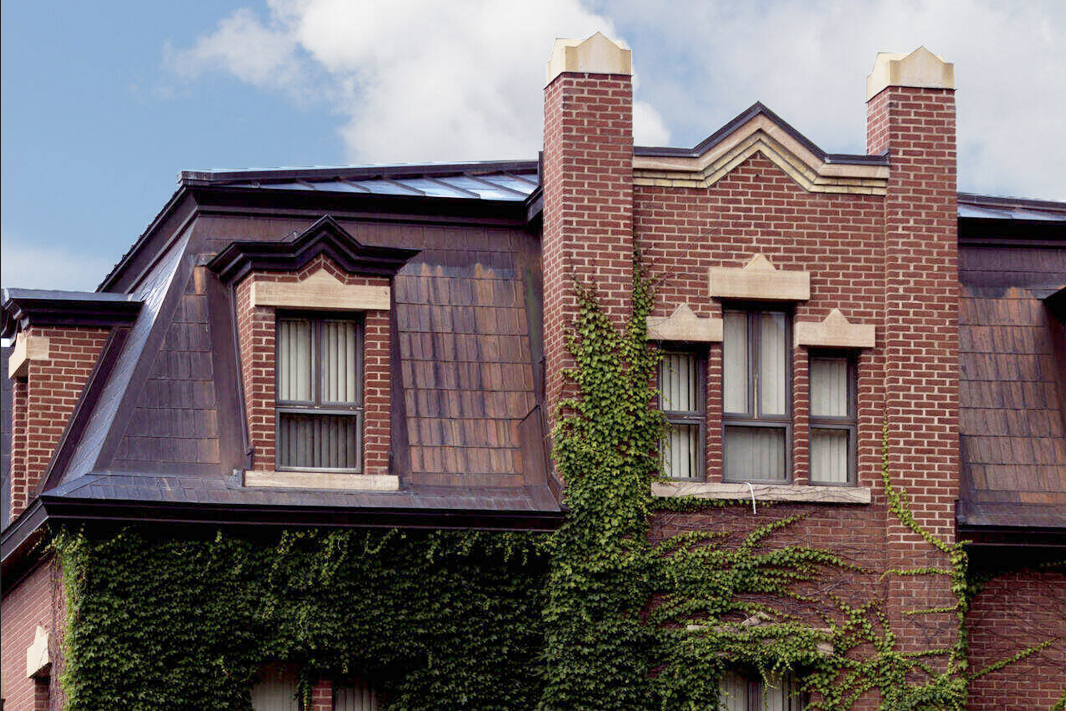 A historic brick building with ivy climbing the facade. It features decorative gables and two chimneys. The slate roof has dormer windows, and the sky is partly cloudy.